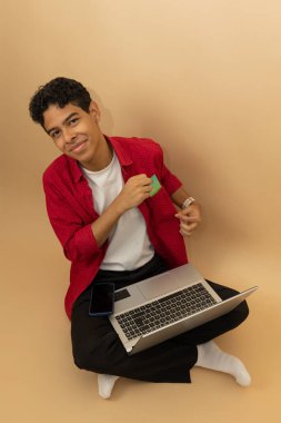 Genç, neşeli, esmer, Latin bir adamın açık kahverengi arka planda bilgisayarından alışveriş yapmak için cebinden bir kart çıkarmasının dikey görüntüsü.