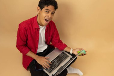 Kahverengi tenli genç bir Latin adamın elinde bir kartla oturmuş açık kahverengi bir arka planda reklam alanı olan bilgisayarından alışveriş edişinin üst görüntüsü.