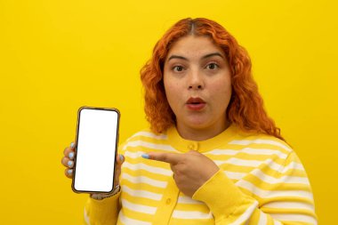 Çilli, kıvırcık saçlı, şaşırmış bir ifadeyle genç bir kadın. Kopyalama alanı olan sarı arka plandaki cep telefonunun boş ekranını işaret ediyor.