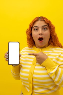 Sarı arka plandaki cep telefonunun boş ekranını gösteren kıvırcık kızıl saçlı, çilli bir kadının dikey görüntüsü.