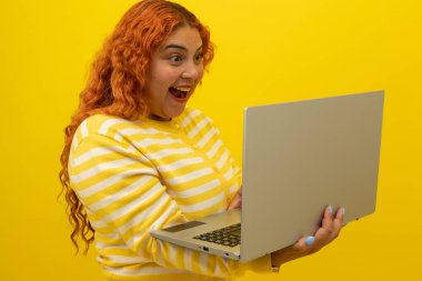 Elinde dizüstü bilgisayar tutan kıvırcık saçlı genç bir kadın telif alanı olan sarı bir arka planda sevinç ifadesi karşısında şaşırdı. Bilgisayarda sürpriz kavramı.