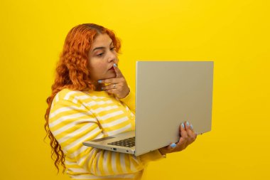 El üstünde el ele tutuşan düşünceli genç kızıl saçlı bir kadın sarı arka planda dizüstü bilgisayar kullanıyor. Bilgisayarda düşünceli olma kavramı.