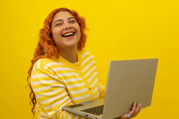 Kırmızı kıvırcık saçlı genç bir kadın, sarı bir arka planda dizüstü bilgisayarda neşeli bir ifade, kopyalama alanı, bilgisayardaki eğlence konsepti