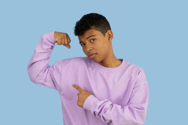 Genç, kahverengi tenli Latin bir adam elleriyle güçlü bir jest yapıyor, leylak rengi bir kazak giyiyor ve açık mavi arka plandaki kaslara işaret ediyor.