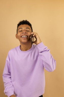 Latin bir gencin eflatun eşofman üstü giyen ve cep telefonuyla mutlu bir şekilde konuşan bir gencin dikey görüntüsü. Kopyalama alanı olan açık kahverengi bir arka planda.