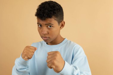 Koyu tenli genç açık mavi bir kazak giyiyor. İki eli havada. Açık kahverengi bir arka planda dövüşmeye hazır.