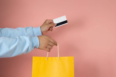 Mavi kazaklı bir adam elinde bir kart ve pembe arka planda sarı bir alışveriş çantası tutuyor.