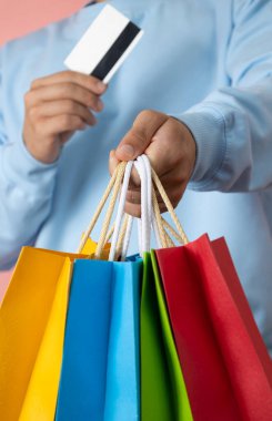 Mavi kazaklı, elinde banka kartı olan ve pembe arka planda çok renkli alışveriş torbaları taşıyan tanınmamış bir adam.