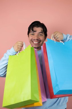 Pembe arka planda çok renkli alışveriş torbaları taşıyan gülümseyen genç adam, alışveriş konsepti
