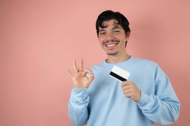 Elinde kredi kartıyla gülümseyen çilli adam pembe arka planda el hareketi yapıyor, kart alışverişi kavramı.