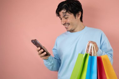 Akıllı telefonunu kullanan neşeli bir adam pembe arka planda çok renkli alışveriş torbaları tutuyor.