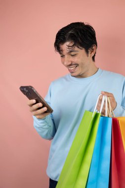 Akıllı telefonunu kullanan neşeli bir adam pembe arka planda çok renkli alışveriş torbaları tutuyor.