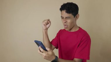 Koyu tenli, kırmızı gömlekli bir adam cep telefonuyla açık kahverengi bir arka planda kutlama yapıyor.