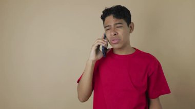 Kırmızı tişörtlü genç Latin adam cep telefonuyla açık kahverengi arka planda konuşuyor, iletişim konsepti.