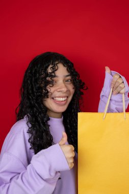Mor kazaklı neşeli genç bir kadın kırmızı arka planda sarı alışveriş torbası tutarken başparmağını kaldırıyor.