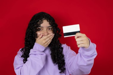 Kırmızı arka planda banka kartı tutan eli ağızda şaşırmış genç bir kadın.