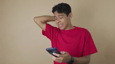 Eli kafasında endişeli genç bir Latin adam açık kahverengi bir arka planda cep telefonuna bakıyor.