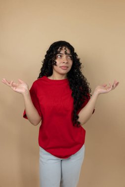 Açık elleri olan genç bir kadın, açık kahverengi bir arka planda, düşünceli ve kafası karışık bir ifadeyle...