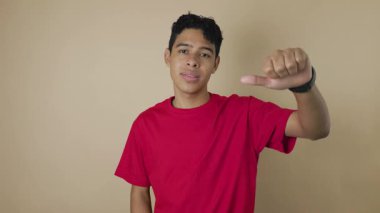 Kırmızı tişörtlü, kahverengi tenli genç bir adam açık kahverengi arka plana başparmak işareti gösteriyor, pazarlama, reddetme veya tatminsizlik için fotokopi alanı olan bir video.