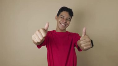 Kırmızı tişörtlü gülümseyen Latin adam açık kahverengi arka plan, başarı, onay, güven ve motivasyon kavramına başparmak işareti yapıyor.
