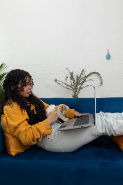 Oturma odasındaki mavi kanepede uzanmış kedisine eşlik eden dizüstü bilgisayarı, uzaktan çalışma ve evdeki modern yaşam tarzı üzerine odaklanmış genç bir kadın.