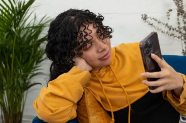 Oturma odasındaki mavi koltukta uzanmış genç bir kadın cep telefonunu tutuyor ve izlemekten zevk alıyor. Dijital bağlantı, rahatlama ve modern ev hayatı kavramı.