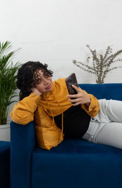 Oturma odasındaki mavi koltukta uzanmış genç bir kadın cep telefonunu tutuyor ve izlemekten zevk alıyor. Dijital bağlantı, rahatlama ve modern ev hayatı kavramı.
