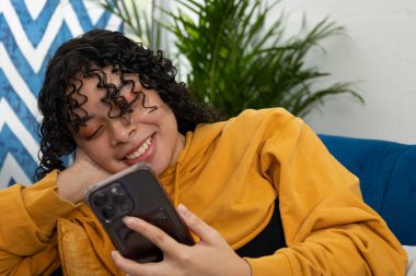 Oturma odasındaki mavi kanepede uzanmış genç bir kadının cep telefonu, mutluluk konsepti, dijital eğlence ve modern ev hayatını kullanırken gülümsemesini yakından çek.