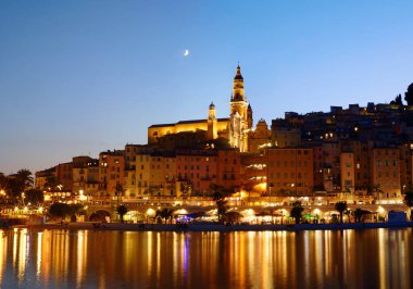 Fransa 'nın Menton kentinin panoramik gece manzarası, aydınlatılmış eski bir kasaba su üzerinde yansıyor, ve Akdeniz kıyısı koyu mavi bir gökyüzünün altında parlıyor.