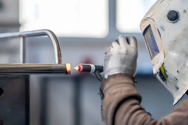An engineer in a welding helmet is using a welding machine to join together a metal pipe, showing skilled gestures in working with aluminium fixture