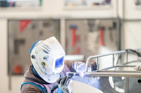 In a factory, a man in sports gear with a welding helmet and personal protective equipment is welding an auto part with an electric blue machine