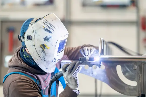 A man in personal protective equipment wearing a welding helmet is using a machine to weld a piece of metal in an engineering factory