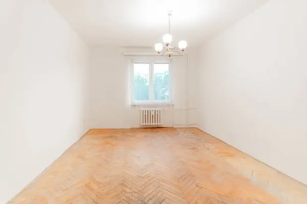 stock image Spacious, well lit room featuring a large window, a chandelier, and old parquet flooring, ready for a complete renovation