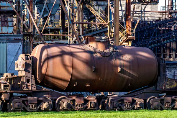 Eski paslı demiryolu tankı bir fabrika binasının önünde endüstriyel kullanımının yıpranmasını ve yıpranmasını gösteriyor. Öğleden sonra güneşi tankın ve etrafındaki çimlerin üzerine gölge düşürüyor.