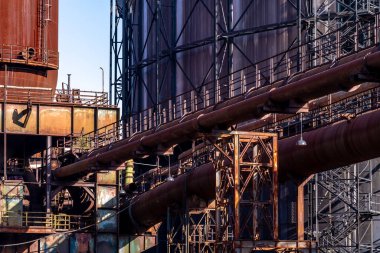 Paslı endüstriyel borular terk edilmiş bir alanda depolama tankları ve çelik yapılar önünde kesişiyor.