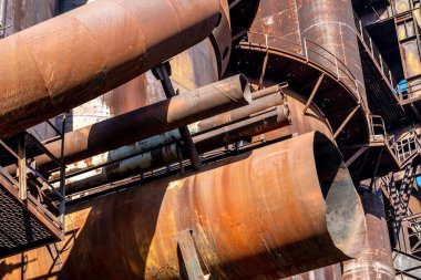 Büyük paslı borular ve metal yapılar görüntüye hakim olup endüstriyel ve soyut bir manzara yaratıyor. Sıcak güneş ışığı eskimiş metalin dokusunu ve detaylarını vurgular.