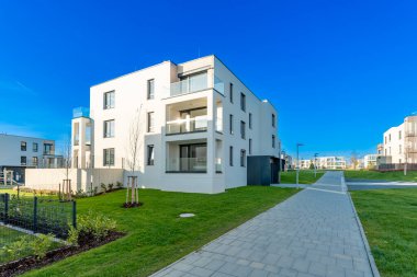 Balkonları ve geniş pencereleri olan, yeşil çimlerle çevrili, sakin bir banliyö mahallesinde, berrak mavi gökyüzünün altında, asfalt bir patikayla çevrili modern beyaz konut binası.