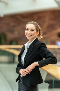 Modern bir ofis binasında korkuluklara yaslanırken gülümseyen neşeli bir iş kadınının portresi, şirket ortamında güven ve profesyonelliği iletiyor.