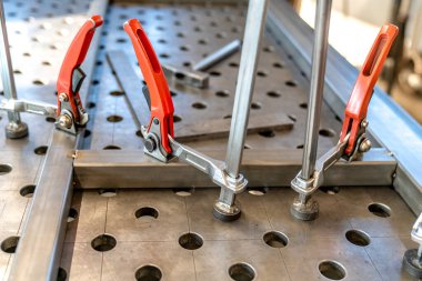 Metal parçaları bir kaynak masasında güvenli bir şekilde bir araya getiren birkaç klemp, yoğun bir metal atölyesinde birleşme sürecine hazırlanıyor, hassasiyet ve zanaatkarlık sergiliyor