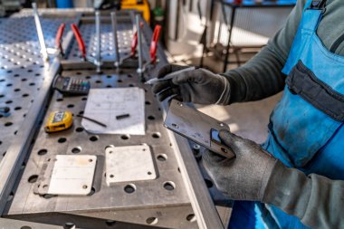 Metal işçisi, atölyedeki metal bir plaka üzerindeki ölçümleri dikkatlice işaretlerken, bir kalem kullanıyor ve arka planda aletler ve ekipmanlarla teknik çizimlere atıfta bulunuyor.