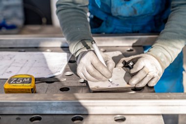 Metal işçisi eldiven takıyor. Metal plaka üzerindeki ölçümleri dikkatle işaretlemek için kalem kullanıyor. Yakınlarda bir metal atölyesinde bant metresi ve planlar var.