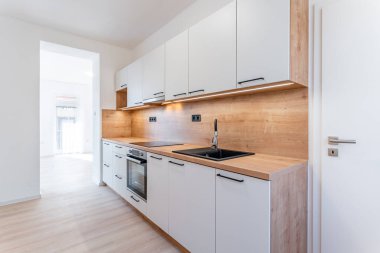 Entegre ekipmanların yer aldığı modern beyaz ve ahşap mutfak, ışıklarla aydınlatılmış, minimalist tasarımı ile parlak ve davetkar bir ev alanı yaratıyor.