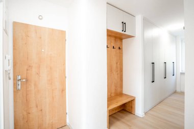 Modern koridor iç dizaynı, ahşap bir kapı, beyaz gardırop, ceket askısı ve küçük bir bank içeriyor, çağdaş bir evde şık ve işlevsel bir giriş yaratıyor.