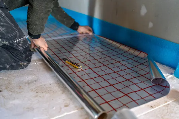 İnşaat işçisi yere alüminyum folyo yalıtımı yerleştiriyor ve termal performans ve enerji verimliliğini artırmak için zemin altı ısıtma sistemi kurulumuna hazırlanıyor.