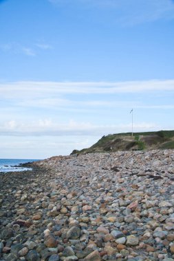 Deniz kıyısındaki taş plajı. Tepesinde Danimarka bayrağı olan bir bayrak direği. Güneşli bir günde gökyüzünde bulutlar olan Danimarka kıyıları. Manzara suya bakıyor.