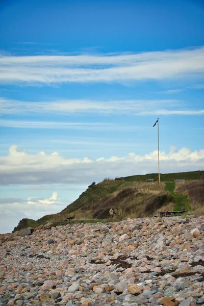 海のそばの石のビーチ 丘の上にデンマーク国旗が掲揚されている 空に雲と晴れた日にデンマークの海岸 水で撮影された風景 — ストック写真