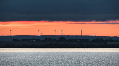 Rüzgar türbini, yenilenebilir enerji, gün batımında bir gölde. İklim değişikliği için temiz elektrik. İskandinavya 'da turuncu güneşli bulutlu bir gökyüzü.