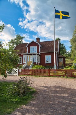 Küçük bir ülkede tipik bir kırmızı-beyaz ev. Beyaz bahçe kapısı, kahverengi çit. İsveç bayrağıyla bayrak direği. Bahçedeki yeşil çimenlik. Arka planda ağaçlar var. Küçük beyaz bulutlu mavi gökyüzü