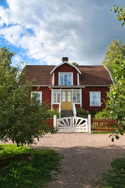 一个典型的红色和白色的瑞典小房子 白色花园大门 棕色栅栏 花园里的绿色草坪 树在前景中 白云小 斯堪的纳维亚景观 — 图库照片