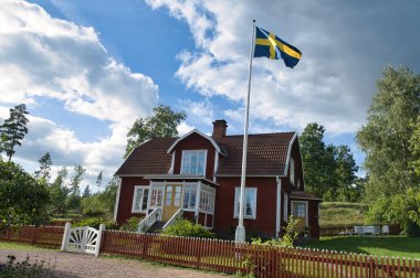 Küçük bir ülkede tipik bir kırmızı-beyaz ev. Beyaz bahçe kapısı, kahverengi çit. İsveç bayrağıyla bayrak direği. Bahçedeki yeşil çimenlik. Arka planda ağaçlar var. Küçük beyaz bulutlu mavi gökyüzü